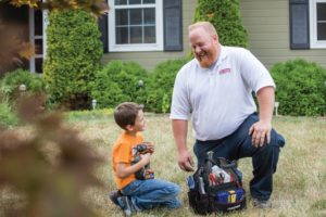 HVAC technician kneeling down in a yard with his tool kit to talk to a child.